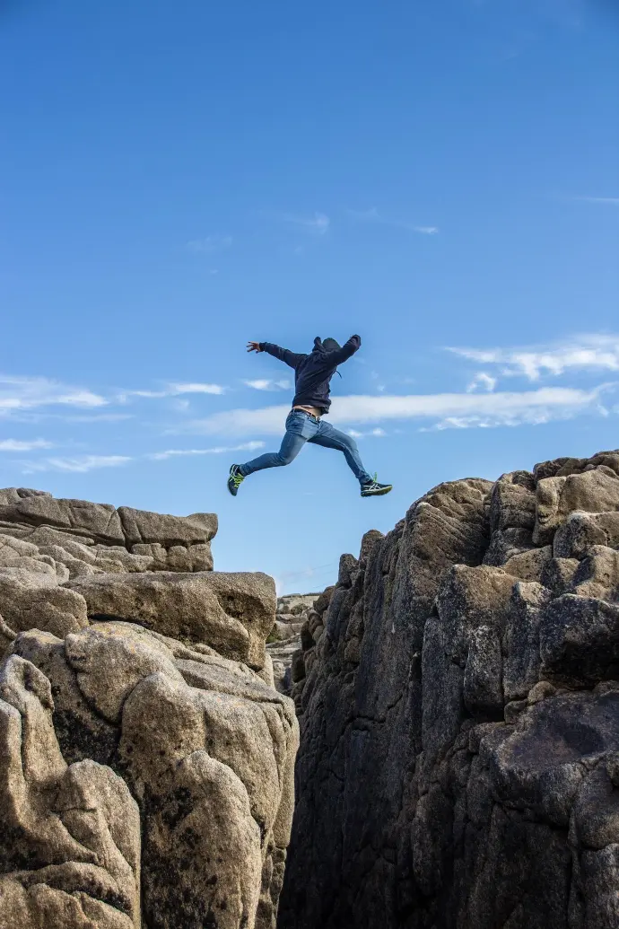 man in black jacket and blue denim jeans jumping on brown rocky mountain during daytime